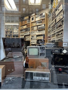 A vintage electronics store display behind a glass window, showcasing a collection of retro audio and video equipment. The shelves are filled with various devices, including old televisions, radios, amplifiers, and turntables. Reflection of the street and pedestrians create an overlay effect on the glass.