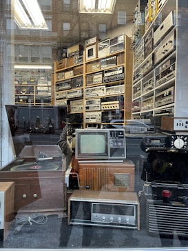 A vintage electronics store display behind a glass window, showcasing a collection of retro audio and video equipment. The shelves are filled with various devices, including old televisions, radios, amplifiers, and turntables. Reflection of the street and pedestrians create an overlay effect on the glass.