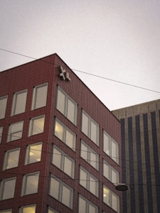 A multi-story red brick building with large windows is viewed from a low angle. The facade is adorned with a prominent white logo near the top corner. Overhead utility lines intersect the sky, and another tall building with vertical lines is partially visible in the background.