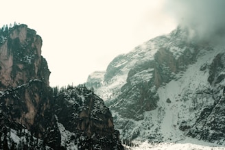 A rugged mountain landscape with steep cliffs covered in patches of snow. Tall dark rocks and evergreen trees create a stark contrast against the lighter snow-covered areas. Mist hovers over the mountain tops, adding a mysterious and serene quality to the scene.
