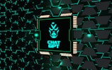 A stylized representation of a computer chip is integrated into a black and teal geometric background. The chip features a minimalist, futuristic face design along with the text 'CHAT GPT' in bold, glowing teal. The background consists of repeating black shapes outlined in teal, creating a high-tech, digital atmosphere.