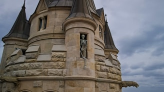 A stone tower with gothic architectural features, including pointed roofs and detailed carvings. A skeleton is positioned prominently in a narrow recess, suggesting a Halloween or eerie theme. The sky is overcast, enhancing the mysterious atmosphere.