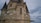A stone tower with gothic architectural features, including pointed roofs and detailed carvings. A skeleton is positioned prominently in a narrow recess, suggesting a Halloween or eerie theme. The sky is overcast, enhancing the mysterious atmosphere.