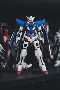 A detailed model of a Gundam robot, featuring a complex design with prominent colors of blue, white, and red. It is positioned upright with intricate mechanical features, standing on a dark surface with a blurred background.