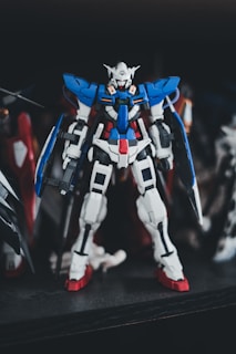 A detailed model of a Gundam robot, featuring a complex design with prominent colors of blue, white, and red. It is positioned upright with intricate mechanical features, standing on a dark surface with a blurred background.