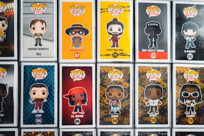 A collection of Pop! Vinyl figures displayed in their boxes, featuring various characters from television, music, and movies. Each box includes a cartoonish depiction of the character on a vibrant background, with their name and product number written below. The figures include characters like Dwight Schrute, Hamburlgar, Post Malone, Eazy-E, and others.