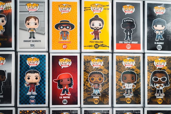A collection of Pop! Vinyl figures displayed in their boxes, featuring various characters from television, music, and movies. Each box includes a cartoonish depiction of the character on a vibrant background, with their name and product number written below. The figures include characters like Dwight Schrute, Hamburlgar, Post Malone, Eazy-E, and others.