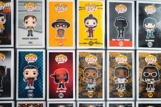 A collection of Pop! Vinyl figures displayed in their boxes, featuring various characters from television, music, and movies. Each box includes a cartoonish depiction of the character on a vibrant background, with their name and product number written below. The figures include characters like Dwight Schrute, Hamburlgar, Post Malone, Eazy-E, and others.