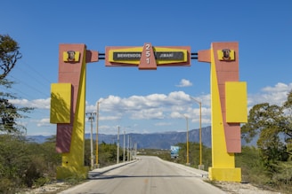 A large, colorful archway spans across a road, welcoming visitors with the text 'Bienvenidos a Jimaní.' The arch features bold geometric designs in yellow and pink, with a clear blue sky and mountainous landscape in the background. Streetlights line the road, and there is sparse vegetation along the sides.
