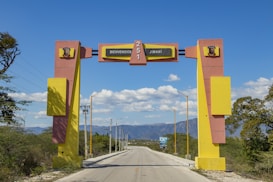 A large, colorful archway spans across a road, welcoming visitors with the text 'Bienvenidos a Jimaní.' The arch features bold geometric designs in yellow and pink, with a clear blue sky and mountainous landscape in the background. Streetlights line the road, and there is sparse vegetation along the sides.
