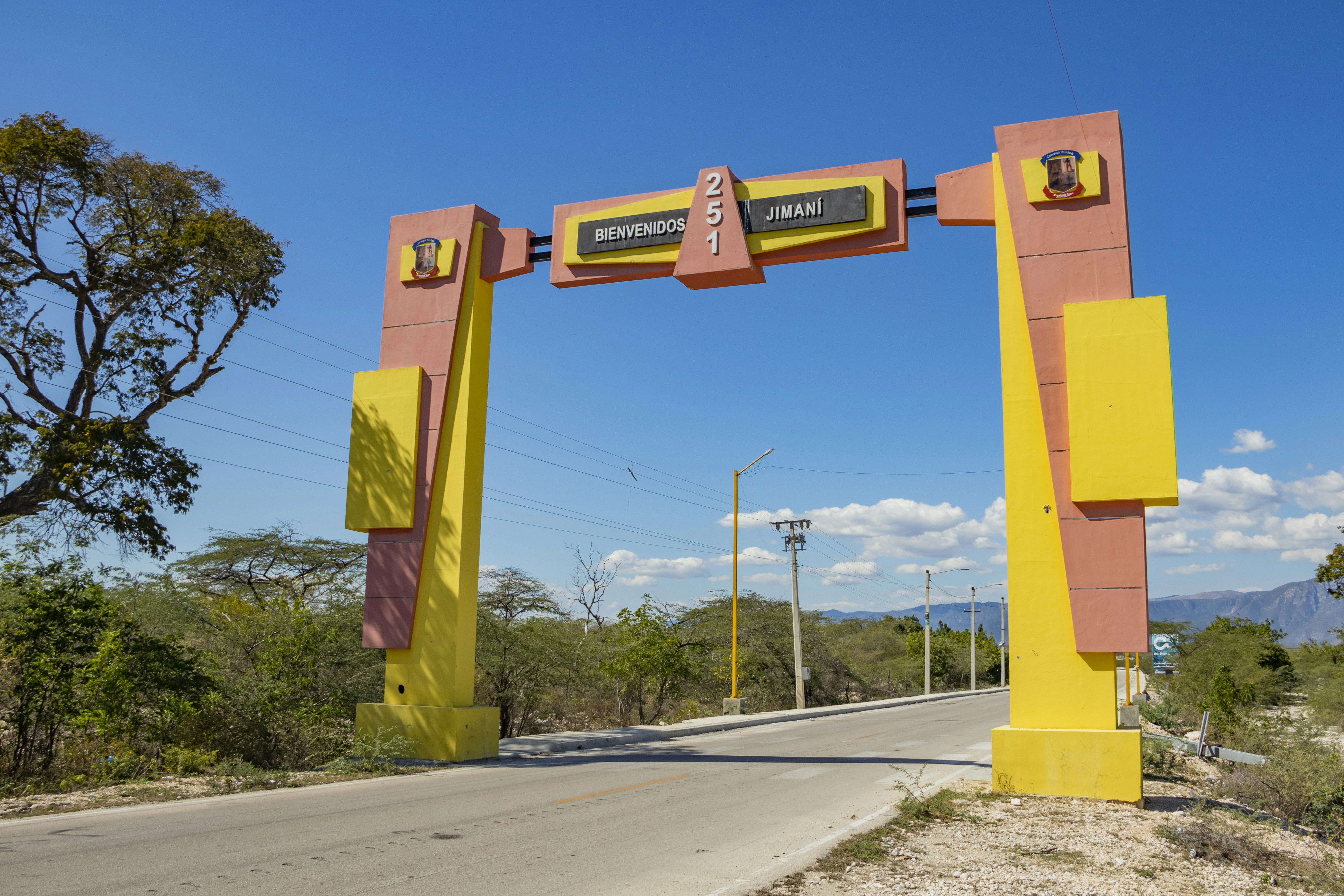 An entrance archway with the words 'Bienvenidos a Jimaní' stands over a road. The structure features bold geometric shapes in yellow and red-orange. Surrounding the road are trees, and in the background, mountains are visible under a clear blue sky.