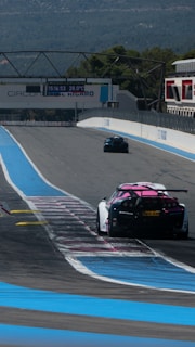 Two racing cars are seen driving on a motor racing track, marked with blue and white lines. The track is wide and mostly empty, surrounded by mountains and trees in the background. A large electronic display shows the time and temperature, mounted above the track.