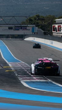 Two racing cars are seen driving on a motor racing track, marked with blue and white lines. The track is wide and mostly empty, surrounded by mountains and trees in the background. A large electronic display shows the time and temperature, mounted above the track.