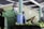 A person in a green uniform and hairnet is wearing a face mask while operating a sprayer. They appear to be working in a facility with green walls and are spraying a row of bananas.