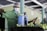 A person in a green uniform and hairnet is wearing a face mask while operating a sprayer. They appear to be working in a facility with green walls and are spraying a row of bananas.