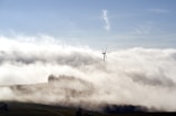 A wind turbine stands tall amidst a landscape blanketed in fog, with the top of the turbine visible above the mist. The surrounding area appears to be hilly and there is a distant silhouette of trees emerging from the fog. The sky is clear and blue, adding a contrast to the misty setting.