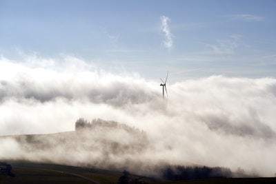 A wind turbine stands tall amidst a landscape blanketed in fog, with the top of the turbine visible above the mist. The surrounding area appears to be hilly and there is a distant silhouette of trees emerging from the fog. The sky is clear and blue, adding a contrast to the misty setting.