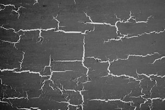 A textured surface with numerous cracks spreading randomly across a dark background. The cracks are white and irregularly shaped, forming a network-like pattern.