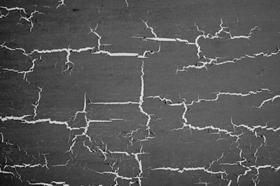 A textured surface with numerous cracks spreading randomly across a dark background. The cracks are white and irregularly shaped, forming a network-like pattern.