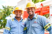 Two individuals wearing blue work shirts and yellow safety helmets are standing side by side, smiling. They are positioned in front of industrial equipment, which is red and white. The setting appears to be outdoors, with trees and a blue sky visible in the background.