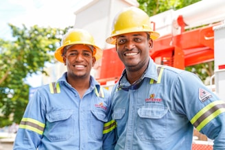 Two individuals wearing blue work shirts and yellow safety helmets are standing side by side, smiling. They are positioned in front of industrial equipment, which is red and white. The setting appears to be outdoors, with trees and a blue sky visible in the background.