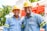 Two individuals wearing blue work shirts and yellow safety helmets are standing side by side, smiling. They are positioned in front of industrial equipment, which is red and white. The setting appears to be outdoors, with trees and a blue sky visible in the background.