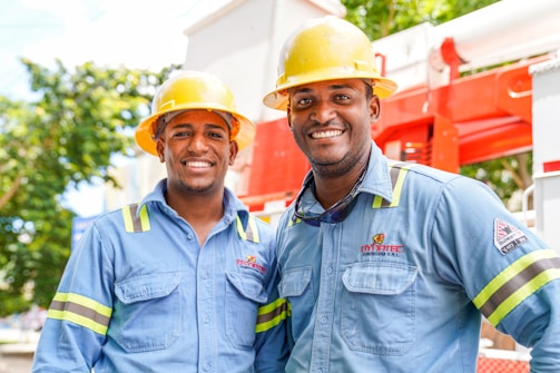 Two individuals wearing blue work shirts and yellow safety helmets are standing side by side, smiling. They are positioned in front of industrial equipment, which is red and white. The setting appears to be outdoors, with trees and a blue sky visible in the background.