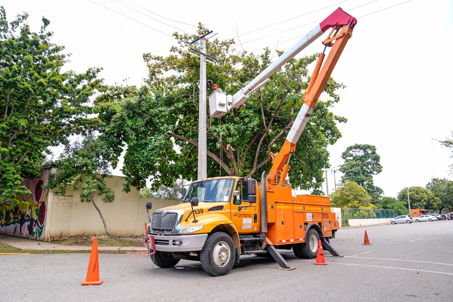 An orange aerial bucket truck with outriggers deployed and its boom raised beside a leafy tree.
