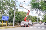 A maintenance crew is working on overhead electrical lines using a white utility truck with a cherry picker. The scene is set on a tree-lined street, and traffic cones are placed around the truck. A street sign indicates directions to various local avenues. The sky is partly cloudy with ample natural light.
