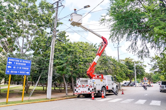 A maintenance crew is working on overhead electrical lines using a white utility truck with a cherry picker. The scene is set on a tree-lined street, and traffic cones are placed around the truck. A street sign indicates directions to various local avenues. The sky is partly cloudy with ample natural light.