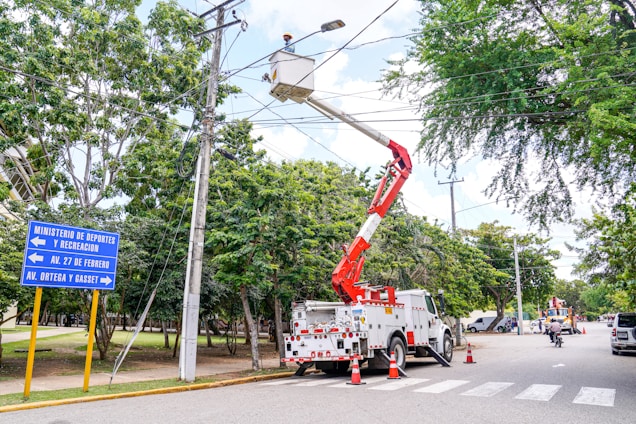 A maintenance crew is working on overhead electrical lines using a white utility truck with a cherry picker. The scene is set on a tree-lined street, and traffic cones are placed around the truck. A street sign indicates directions to various local avenues. The sky is partly cloudy with ample natural light.