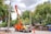 A utility truck with an elevated platform is parked on the side of the street. The platform is raised, and a worker is performing maintenance on overhead cables connected to utility poles. Several people are walking on the sidewalk nearby, and there are lush green trees and a partly cloudy sky in the background. An orange traffic cone is placed in front of the truck.