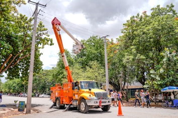 A utility truck with an elevated platform is parked on the side of the street. The platform is raised, and a worker is performing maintenance on overhead cables connected to utility poles. Several people are walking on the sidewalk nearby, and there are lush green trees and a partly cloudy sky in the background. An orange traffic cone is placed in front of the truck.