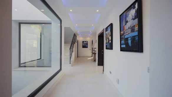 A modern and minimalistic hallway with white walls and a glossy floor. Large framed artworks decorate the walls on the right side, while a glass panel on the left provides a view into another room. Soft recessed ceiling lights create a calm and inviting atmosphere. At the end of the corridor, a staircase ascends, adding depth to the space.