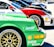 Several sports cars are lined up in a row, showcasing vibrant colors and sponsor logos. The forefront displays a green car with a detailed wheel rim and various brand decals, including Bosch and Shell. Each car features sleek designs, contributing to a dynamic and competitive atmosphere.
