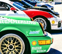Several sports cars are lined up in a row, showcasing vibrant colors and sponsor logos. The forefront displays a green car with a detailed wheel rim and various brand decals, including Bosch and Shell. Each car features sleek designs, contributing to a dynamic and competitive atmosphere.