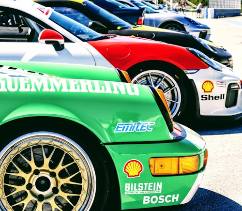Several sports cars are lined up in a row, showcasing vibrant colors and sponsor logos. The forefront displays a green car with a detailed wheel rim and various brand decals, including Bosch and Shell. Each car features sleek designs, contributing to a dynamic and competitive atmosphere.