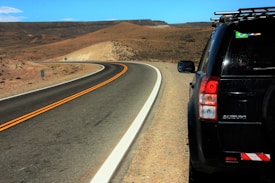 A winding road stretches into a desert landscape under a clear blue sky. A black SUV is parked on the right, partially visible with a Brazilian flag sticker on the window. The surrounding environment is arid, with brown and tan hills.
