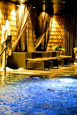 An indoor pool area with water illuminated in blue. The room features elegant and dark-colored drapery hanging from the ceiling, offering a luxurious and serene environment. There are a couple of seating arrangements with dark-toned sofas, adding to the comfort and relaxation ambiance. Towels are draped over a railing, indicating a space for leisure and relaxation. Subtle patterns and plants contribute to the warm atmosphere.