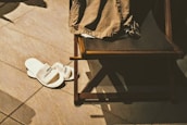 A pair of white hotel slippers is placed on a tiled floor next to a wooden chair with a piece of brown clothing draped over it. Shadows cast by the objects indicate a light source from above.