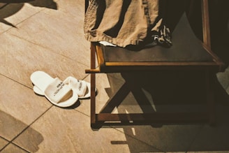 A pair of white hotel slippers is placed on a tiled floor next to a wooden chair with a piece of brown clothing draped over it. Shadows cast by the objects indicate a light source from above.