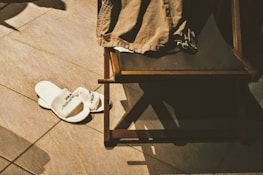 A pair of white hotel slippers is placed on a tiled floor next to a wooden chair with a piece of brown clothing draped over it. Shadows cast by the objects indicate a light source from above.