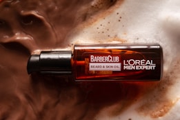 A bottle of L'Oréal Men Expert BarberClub beard and skin oil rests on a surface covered in lather or foamy liquid, suggesting a grooming or skincare setting. The bottle is dark amber with a black dispenser cap.