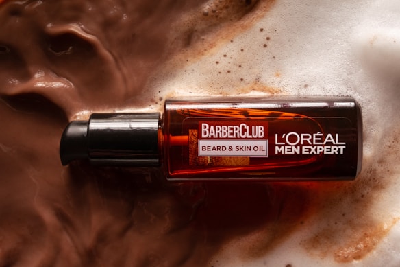 A bottle of L'Or&eacute;al Men Expert BarberClub beard and skin oil rests on a surface covered in lather or foamy liquid, suggesting a grooming or skincare setting. The bottle is dark amber with a black dispenser cap.