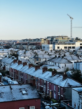 Rows of residential houses with snow-covered roofs are seen in an urban setting. The buildings are of brick construction with chimney stacks. In the background, modern office buildings and a construction crane are visible under a clear sky.