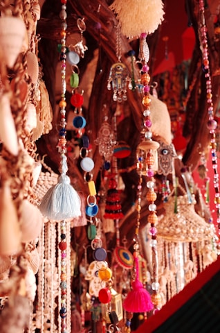 An array of vibrant, colorful decorative beads and charms hang in multiple strands. These include a variety of shapes, such as circular beads, tassels, and ornate ornaments like hamsa hand keychains. The objects are artfully arranged against a wooden backdrop, creating a rich texture with the intricate details of each piece.