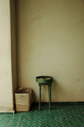 A simple corner of a room featuring a small dark green round table with slender legs and a rectangular wicker basket on a green tiled floor with a chevron pattern. The walls are light beige, and the scene has an understated, minimalist aesthetic.