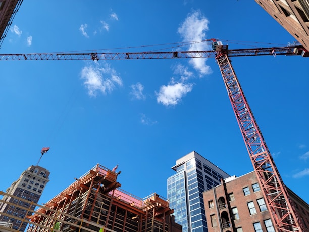 A construction site with a large red crane towering over partially constructed buildings. The scene features several high-rise buildings under a bright blue sky with scattered clouds.