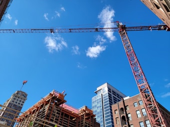 A construction site with a large red crane towering over partially constructed buildings. The scene features several high-rise buildings under a bright blue sky with scattered clouds.