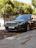 A sleek black luxury sedan is parked on a city street with green leafy trees in the background. The car is angled slightly away from the curb, and dry leaves are scattered on the ground around it. The vehicle features a distinctive front grille and has a European license plate.